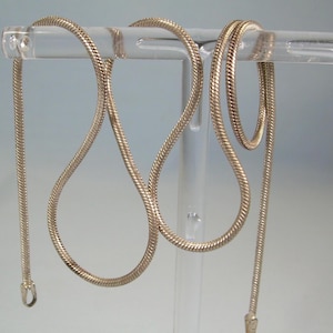 May include: A gold-colored snake chain necklace displayed on a clear acrylic stand. The chain has a smooth, rounded texture and hangs in a looping pattern. The background is a soft, gradient blue.