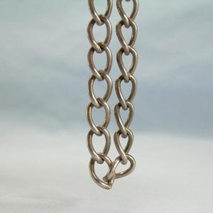 May include: A silver-coloured metal chain featuring elongated, teardrop-shaped links arranged in two parallel vertical strands. The links have a smooth, polished finish and are connected in a continuous, repeating pattern throughout the length of the chain.