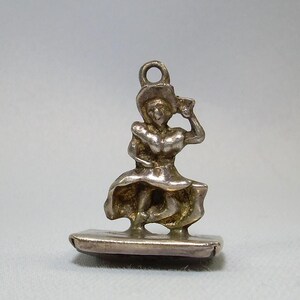 May include: Silver charm of a woman in a dress and hat, standing on a rectangular base.
