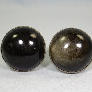 May include: Pair of round, polished obsidian earrings with silver-colored metal trim. One earring is a deep black, while the other has a golden sheen. The earrings are set against a neutral background.