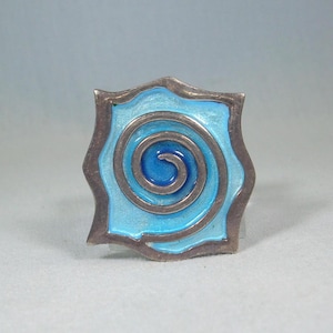 May include: A silver ring with a blue enamel design. The design is a spiral with concentric circles.