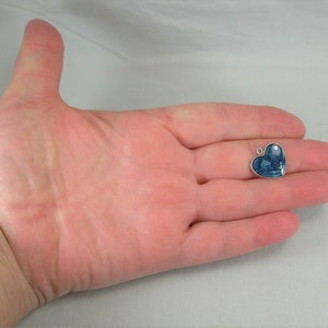 May include: A silver heart-shaped charm with a blue and white image inside. The charm is being held in a person's hand.