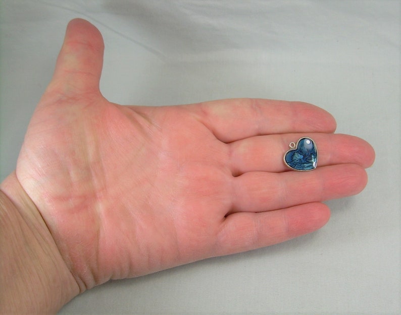 May include: A silver heart-shaped charm with a blue and white image inside. The charm is being held in a person's hand.