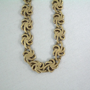 May include: A gold-toned necklace with a repeating floral design. The necklace is composed of interconnected, stylized flower-shaped links. The links are arranged in a symmetrical pattern, creating a continuous chain. The necklace is displayed against a white background.