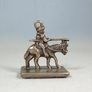Puede incluir: Un charm plateado que representa a un jinete a caballo, sosteniendo un objeto largo. El jinete lleva un sombrero y está sentado sobre el caballo, que se encuentra sobre una base rectangular. El charm tiene un lazo para sujetar.