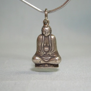 May include: Silver pendant featuring a detailed depiction of a seated Buddha figure.