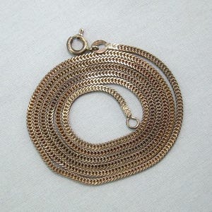 May include: A gold-toned metal chain necklace featuring a tightly woven, flat herringbone or wheat-style link pattern. The necklace is coiled in a circular shape and includes a small, round spring-ring clasp mechanism at the top end of the chain.