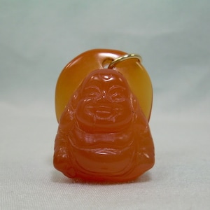 May include: A carved orange-red Buddha pendant with a gold-colored loop for hanging. The Buddha figure is smiling and has a rounded belly. The pendant is set against a larger, oval-shaped orange-yellow background.