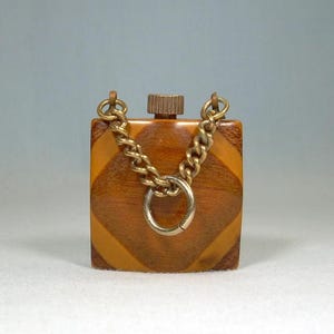 May include: A small, square vintage perfume bottle with a geometric pattern in shades of brown and tan. It features a gold-toned chain handle and a metal screw-on cap. The bottle is likely made of a durable material, and the design suggests a classic aesthetic.