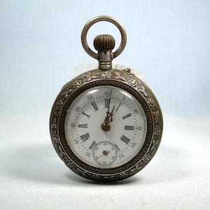 Puede incluir: Un reloj de bolsillo plateado con esfera blanca y números romanos negros. El reloj tiene un diseño floral decorativo alrededor del bisel y un pequeño dial de segundos. El reloj mide aproximadamente 5 cm de diámetro.
