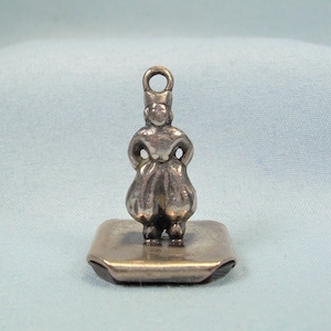 Puede incluir: Encanto de plata de una mujer con un vestido de pie sobre una base cuadrada.