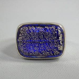May include: A silver ring with a blue glass stone set in the center. The glass has a speckled, silver-like pattern throughout.