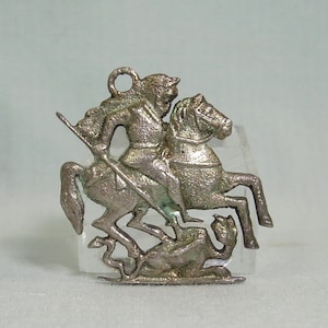 Puede incluir: Pendiente de plata que representa a un caballero a caballo matando a un dragón. El pendiente es detallado e intrincado, con una superficie texturizada.