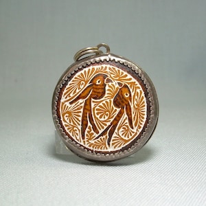 May include: A silver pendant with a round, white enamel design featuring two brown birds with intricate details. The pendant has a silver loop for attaching a chain.