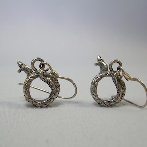 Puede incluir: Un par de pendientes de serpiente de plata con un acabado texturizado. Los pendientes tienen forma de serpiente enroscada en un círculo. La cabeza de la serpiente está mirando hacia adelante y la cola está enroscada alrededor del círculo. Los pendientes cuelgan de un gancho de plata.