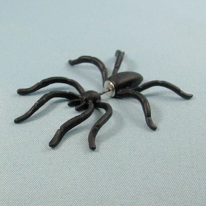 Könnte beinhalten: Schwarzer Spinnenohrring mit einem silbernen Stift. Die Spinne hat acht Beine und einen detaillierten Körper.
