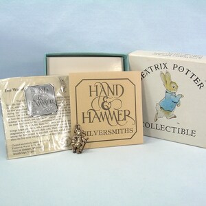 May include: A silver Beatrix Potter charm featuring a rabbit, accompanied by a Hand & Hammer Silversmiths informational card, a protective plastic sleeve with a silver logo sticker, and a white cardboard box with a Peter Rabbit illustration.