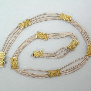 May include: A gold-colored necklace with multiple strands and decorative rectangular clasps. The necklace is made of thin, beige cords and features geometric designs on the clasps. The clasp is closed.