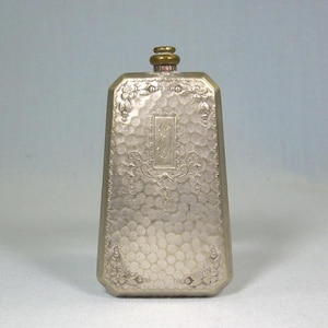 Op de afbeelding: Een zilverkleurige, rechthoekige fles met een getextureerd oppervlak en bloemversieringen. De fles heeft een klein, rechthoekig middenpaneel met een sierlijk ontwerp. De bovenkant heeft een messingkleurige dop. De fles is waarschijnlijk vintage.