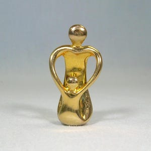 May include: A gold-colored sculpture depicting a parent figure embracing a child. The parent's arms form a heart shape around the child. The sculpture has a smooth, polished finish and is set against a light blue and white background.