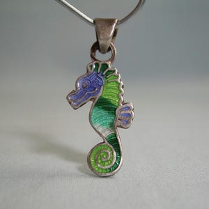 May include: A silver seahorse pendant with green and blue enamel. The seahorse is facing to the right and has a curled tail.