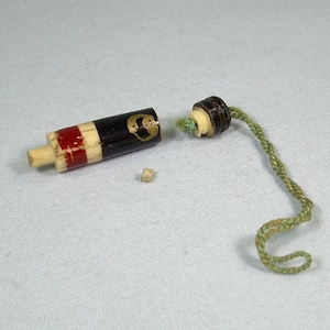 Puede incluir: Un pequeño recipiente de madera desmontado con un diseño negro y rojo, un pequeño dado y un cordón verde. El recipiente tiene un emblema dorado y está hecho de varios materiales.