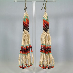 Op de afbeelding: Een paar kralen oorbellen met een crèmekleurige basis en accenten van oranje, rood, bruin en turquoise. De oorbellen hebben een lang, bungelend ontwerp met meerdere strengen kralen.