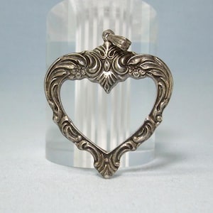 May include: A silver-toned heart-shaped pendant featuring an open center and intricate embossed scrollwork patterns along the frame. The metal surface displays a polished finish with darkened recessed areas to highlight the detailed relief design.