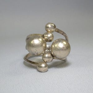May include: A silver ring with a unique design featuring multiple round spheres connected by a thin band.
