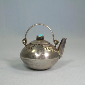 Op de afbeelding: Kleine, zilverkleurige theepot met een afgeronde body, een gebogen tuit en een handvat. Het deksel is versierd met een kleine, ronde turkooizen steen. De theepot heeft geëtste ontwerpen rond de body en het deksel.