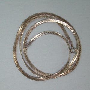 May include: A gold-toned herringbone chain necklace featuring a flat, flexible link design with a polished metallic finish. The necklace is arranged in a circular coil and includes a small, round spring ring clasp mechanism at the end of the chain.