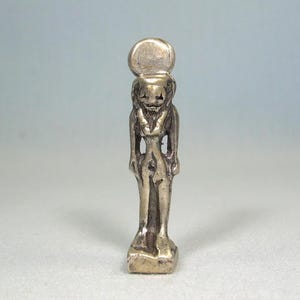 Może przedstawiać: Mała, srebrna statuetka egipskiego bóstwa. Postać ma ludzkie ciało z głową lwa i tarczą słoneczną na szczycie. Statua stoi na prostokątnej podstawie.