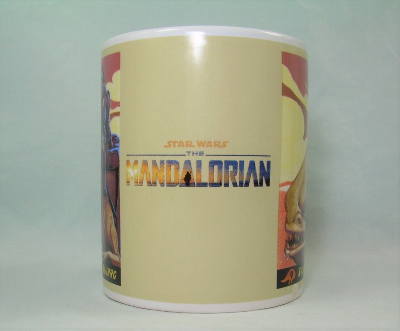 Puede incluir: Una taza de cer&aacute;mica blanca con un fondo beige que presenta el logotipo de Star Wars: The Mandalorian. El logotipo est&aacute; en un esquema de color amarillo, naranja y azul. La taza tiene un gr&aacute;fico de un personaje del programa en el lado izquierdo y un gr&aacute;fico de una criatura del programa en el lado derecho.
