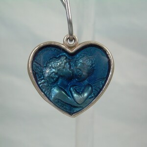 May include: A silver heart-shaped pendant with a blue enamel design of two angels kissing. The pendant has a loop at the top for attaching a chain.