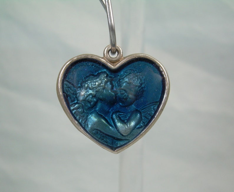 May include: A silver heart-shaped pendant with a blue enamel design of two angels kissing. The pendant has a loop at the top for attaching a chain.