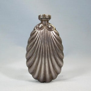 May include: A silver-colored, antique perfume bottle with a shell-like design. The bottle has a small, screw-on cap and a textured, ridged surface. The overall shape is oval, and the bottle appears to be made of metal.