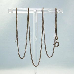 May include: A bronze-toned necklace featuring a fine box chain. The necklace is presented on a clear acrylic stand, highlighting the detailed links and clasp. The chain is a classic design, suitable for pendants or to be worn on its own.