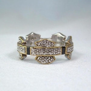 Puede incluir: Un anillo plateado y dorado con un diseño en forma de escudo. El anillo presenta un emblema central con un borde dorado y está incrustado con pequeñas piedras brillantes. Se ven detalles en negro.