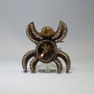 May include: A silver brooch in a bug design. It has a large, oval brown gemstone at the centre, framed by a twisted rope-like border. A smaller, round yellow gemstone sits above the centre stone. The brooch is a unique piece of jewellery.