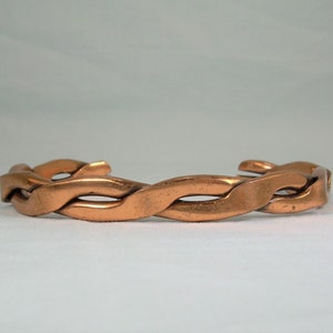 Könnte beinhalten: Ein geflochtenes Kupferarmband. Das Armband besteht aus einem einzigen Stück Kupfer, das verdreht und geflochten ist, um ein einzigartiges und stilvolles Design zu schaffen.