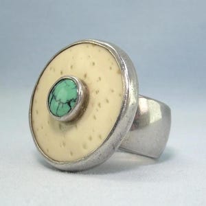 May include: A silver ring with a large, round, off-white face featuring a small, round, turquoise stone in the center. The ring has a thick band and a textured surface on the face.