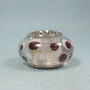 May include: A clear glass bead with a silver metal band and a white and brown polka dot pattern. The bead is marked "Pandora" on the metal band.