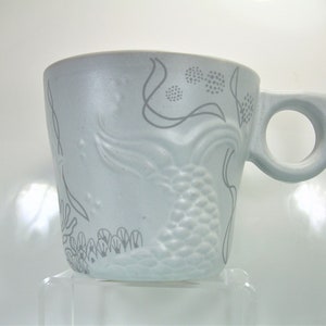 May include: A white ceramic mug featuring a grey mermaid design. The mermaid is surrounded by seaweed and bubbles.