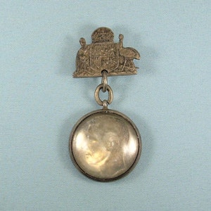 Puede incluir: Un colgante de plata con un camafeo redondo de vidrio transparente que presenta un retrato de perfil. El colgante está unido a una cadena con una pieza superior decorativa que presenta una corona y tres figuras.