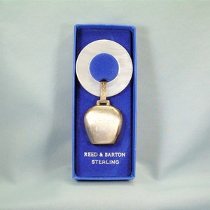 May include: A silver baby rattle in a blue box with a white and blue teething ring. The rattle is shaped like a bell and has the words "Reed & Barton Sterling" on it.