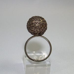 Puede incluir: Un anillo de plata con una gran esfera redonda de malla de alambre en la parte superior.