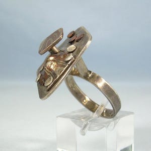 May include: A silver-toned metal ring featuring a large, flat, rectangular top plate. The surface is adorned with several small, circular, copper-colored and silver-toned accents, along with a raised, angled, rectangular piece attached to the top.