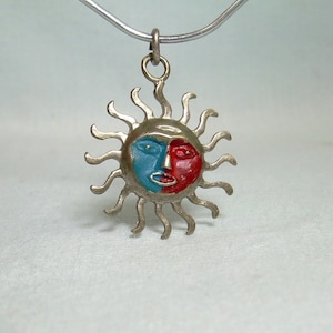 Könnte beinhalten: Silberfarbene Halskette mit einem Anhänger in Form einer Sonne mit Gesicht. Das Gesicht der Sonne ist blau und rot gefärbt. Der Anhänger hängt an einer dünnen, silberfarbenen Kette. Die Sonnenstrahlen sind wellenförmig.