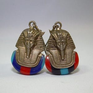 May include: Pair of silver-toned earrings featuring the bust of a pharaoh with a striped headdress. The lower portion of each earring is a half-circle with red, blue, and black stripes. The earrings have a hook closure.