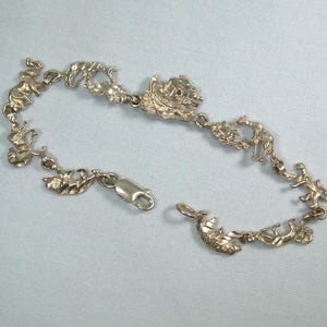 May include: A silver-toned charm bracelet featuring various animal figures, including elephants, giraffes, and other wildlife. The bracelet is unclasped, lying on a light blue surface, showcasing its intricate design and details.